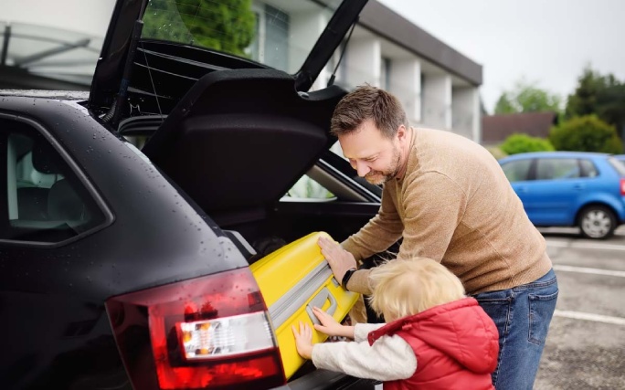 Pai põe uma bagagem na mala de um carro alugado ao lado do seu filho pequeno.