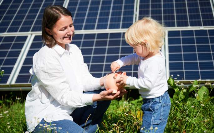 Mãe ajuda filho pequeno a encher porquinho-mealheiro num ambiente de sustentabilidade ecológica formado por painéis solares.