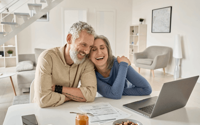 Um casal feliz dentro de casa, sentado à mesa, com papeis e um computador.