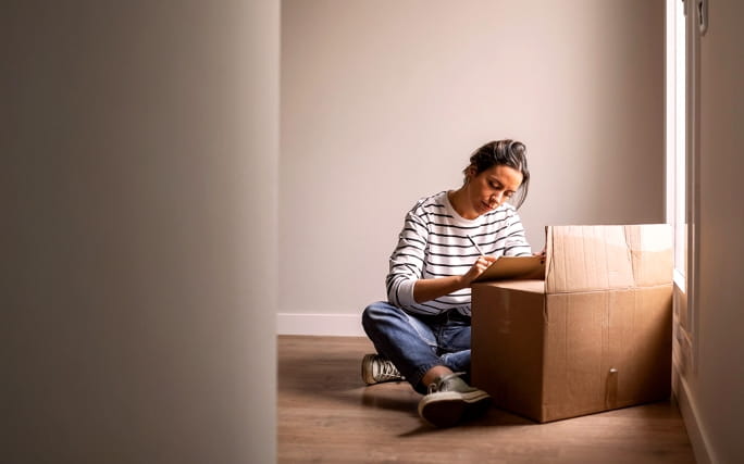 Jovem sentada no chão a escrever numa caixa de cartão durante uma mudança.