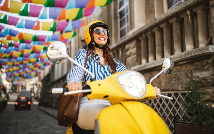 Mulher jovem sorridente, com com carta B, a conduzir uma mota 125 cc pela cidade.
