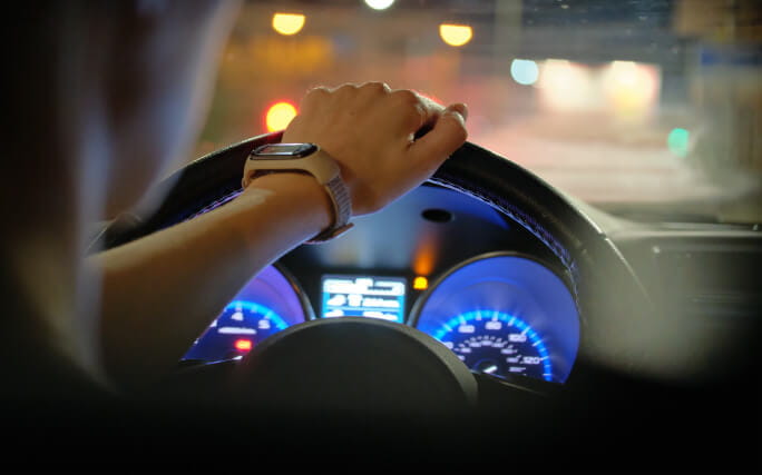 Mãos sobre o volante de um carro em marcha na estrada, sendo visíveis as luzes do painel do carro.  