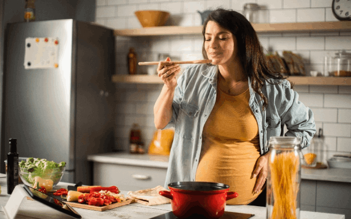Uma mulher jovem em fase de gravidez avançada prova a refeição saudável que está a cozinhar na sua cozinha.