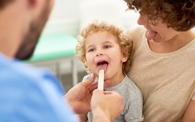 Um médico pediatra examina a boca e garganta de uma criança sentada no colo da mãe.