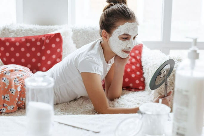 Mulher jovem com máscara de cosmético sustentável aplicada no rosto, olha-se ao espelho  