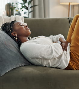 Mulher com expressão de desconforto, deitada num sofá em casa agarrada à barriga por sofrer de dores de estômago.