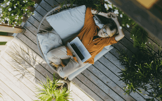 Mulher jovem sentada no chão, num pátio rodeado de plantas, trabalha no seu laptop.