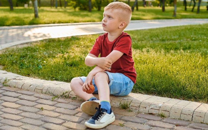 Menino sentado no chão de um jardim agarra o seu cotovelo, devido à dor causada por uma pronação dolorosa.
