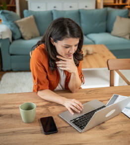 Mulher na sala de casa, sentada à mesa usa um computador portátil.  
