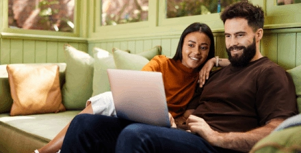Casal sorridente faz um seguro de vida online, sentado no sofá com portátil nos joelhos